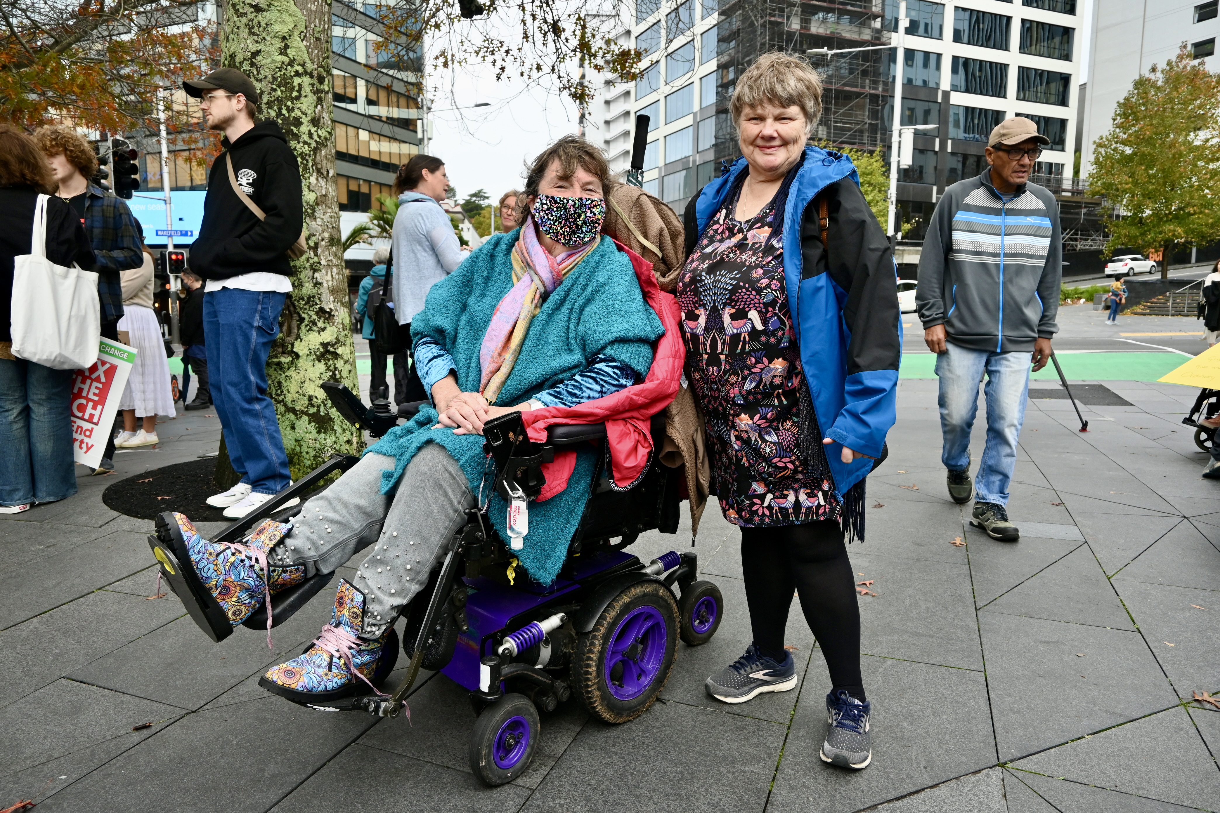 Two disabled women pose for a photo on Queen Street; one is using a powered-wheelchair and wears a face mask. 