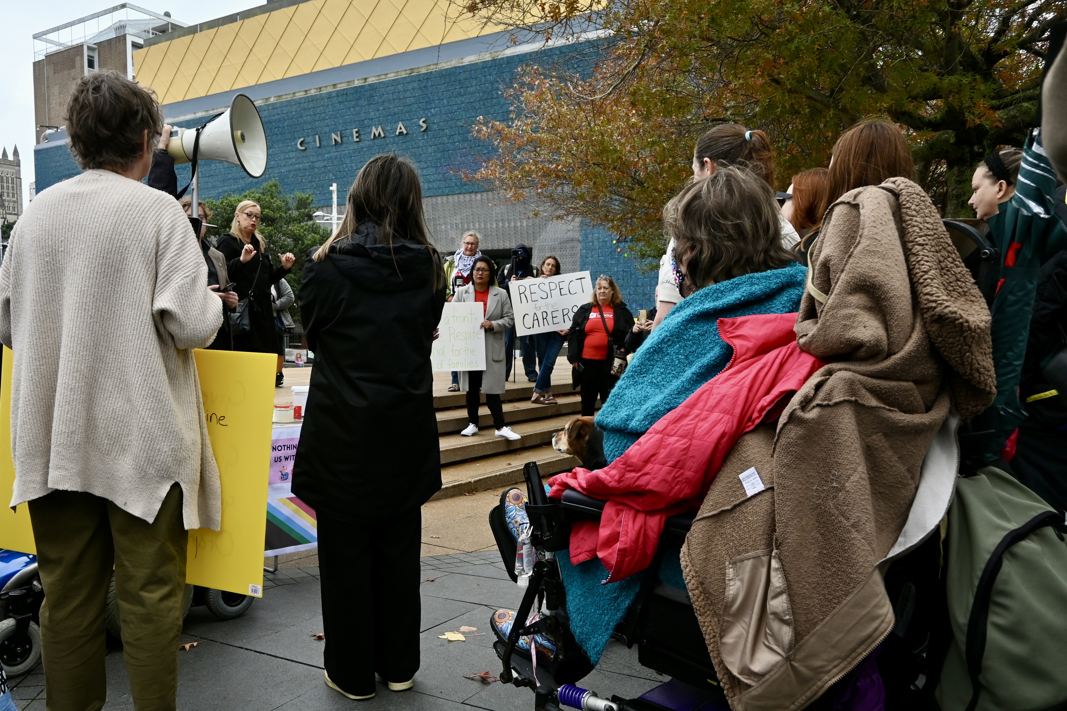 A crowd of protestors listen to speakers at a rally. A sign reads: Respect carers.