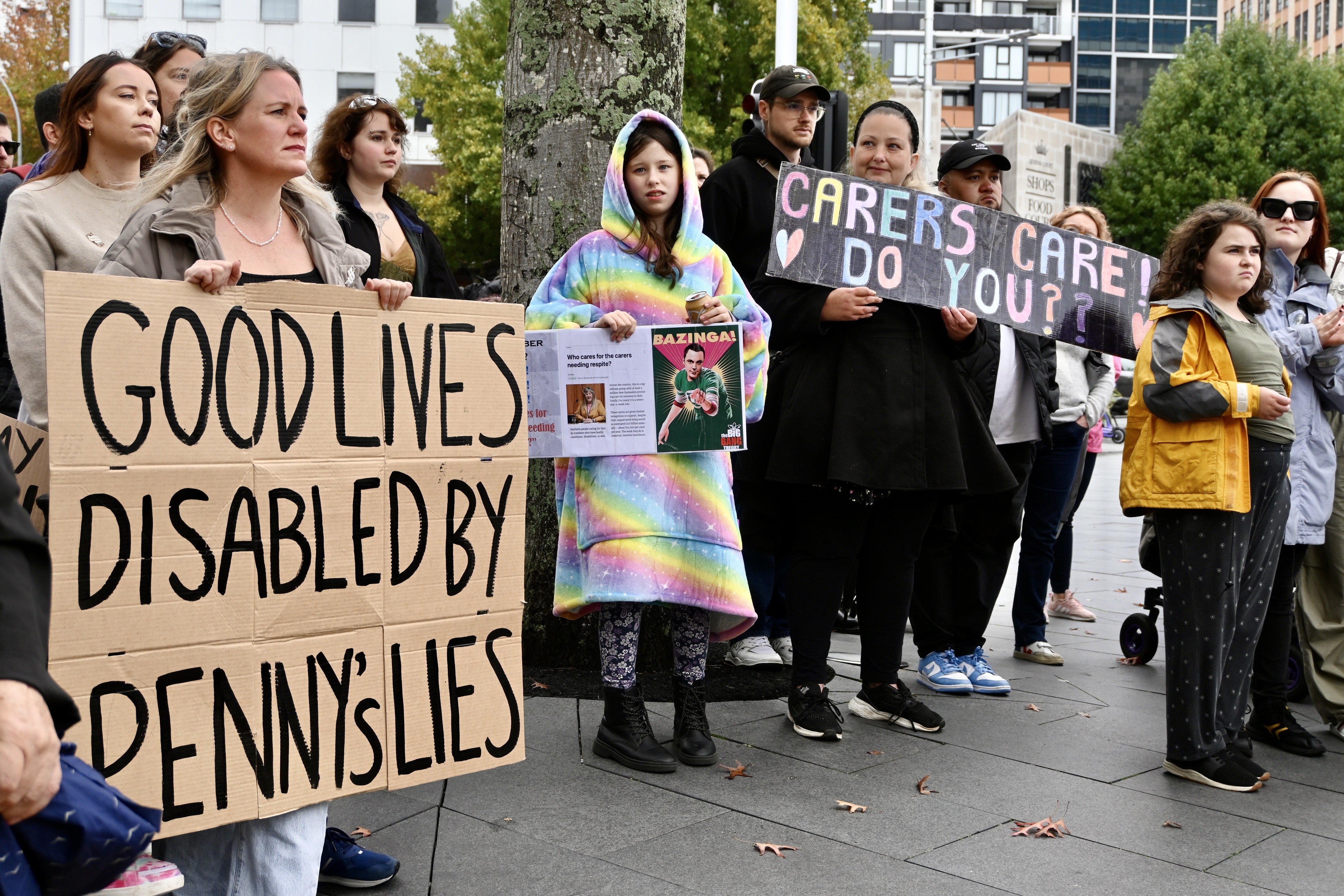 A group of people protest. A cardboard sign reads: Good lives disabled by Penny's lies. Another sign reads: Carers care, do you?
