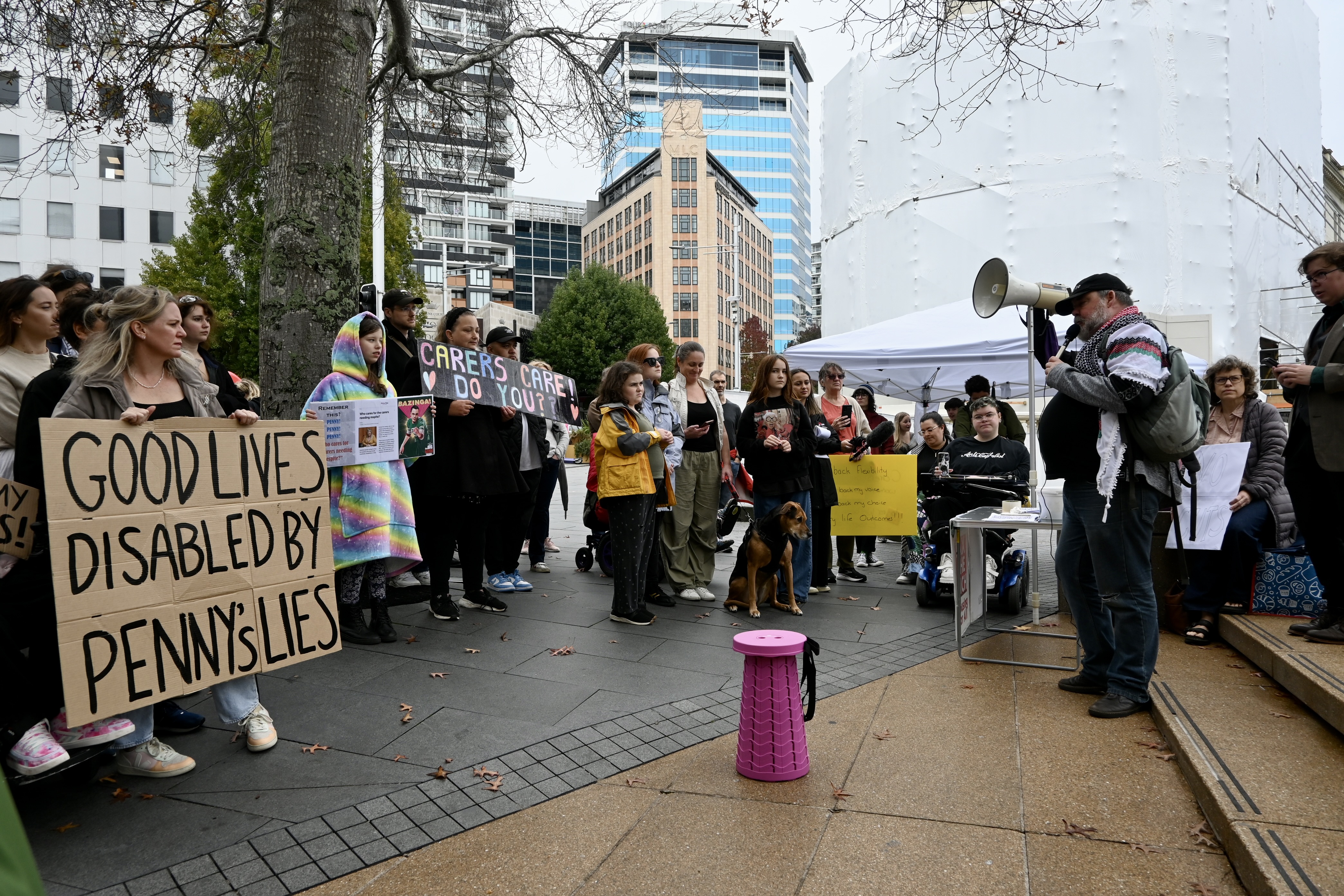 A group of disabled and nondisabled people protest, with a man speaking into a megaphone in front of the crowd. A cardboard sign reads: Good lives disabled by Penny's lies.  Another sign reads: Carers care, do you?