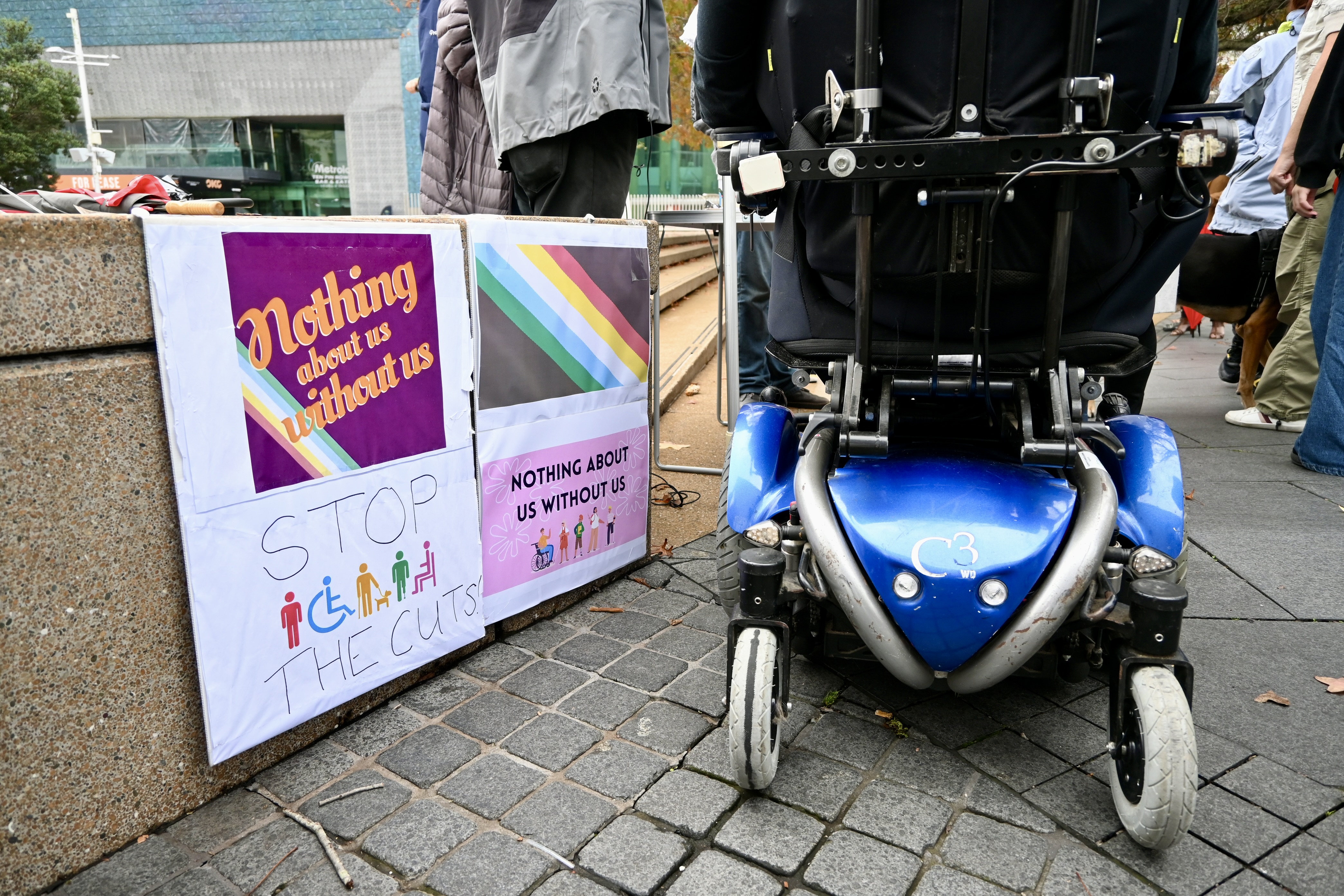 The wheels of a powered-wheelchair are rolling next to signs that read: Nothing about us without us, and Stop the cuts.