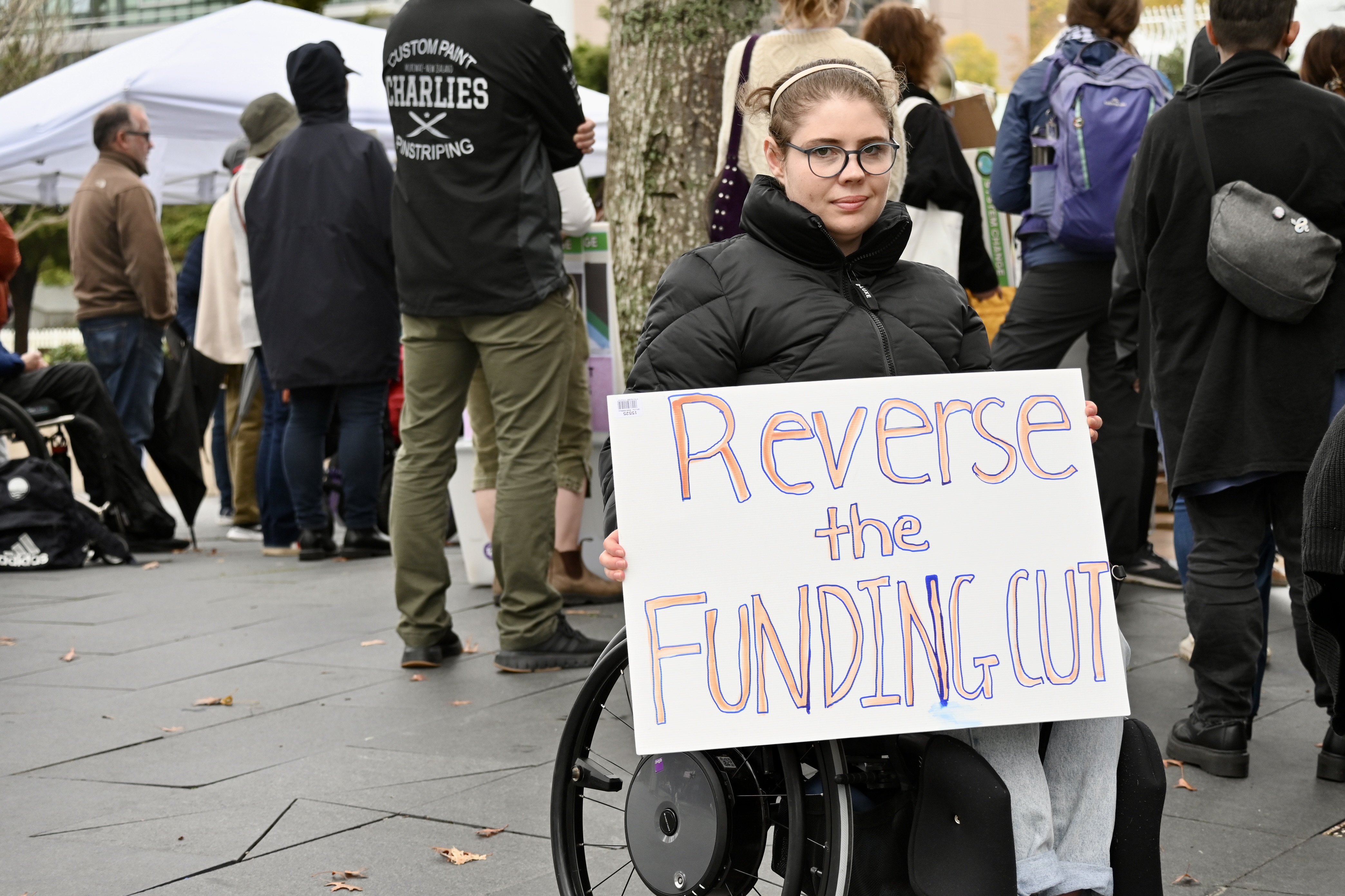 A young wheelchair-user wears glasses and holds up a sign that reads: Revere the funding cut. 