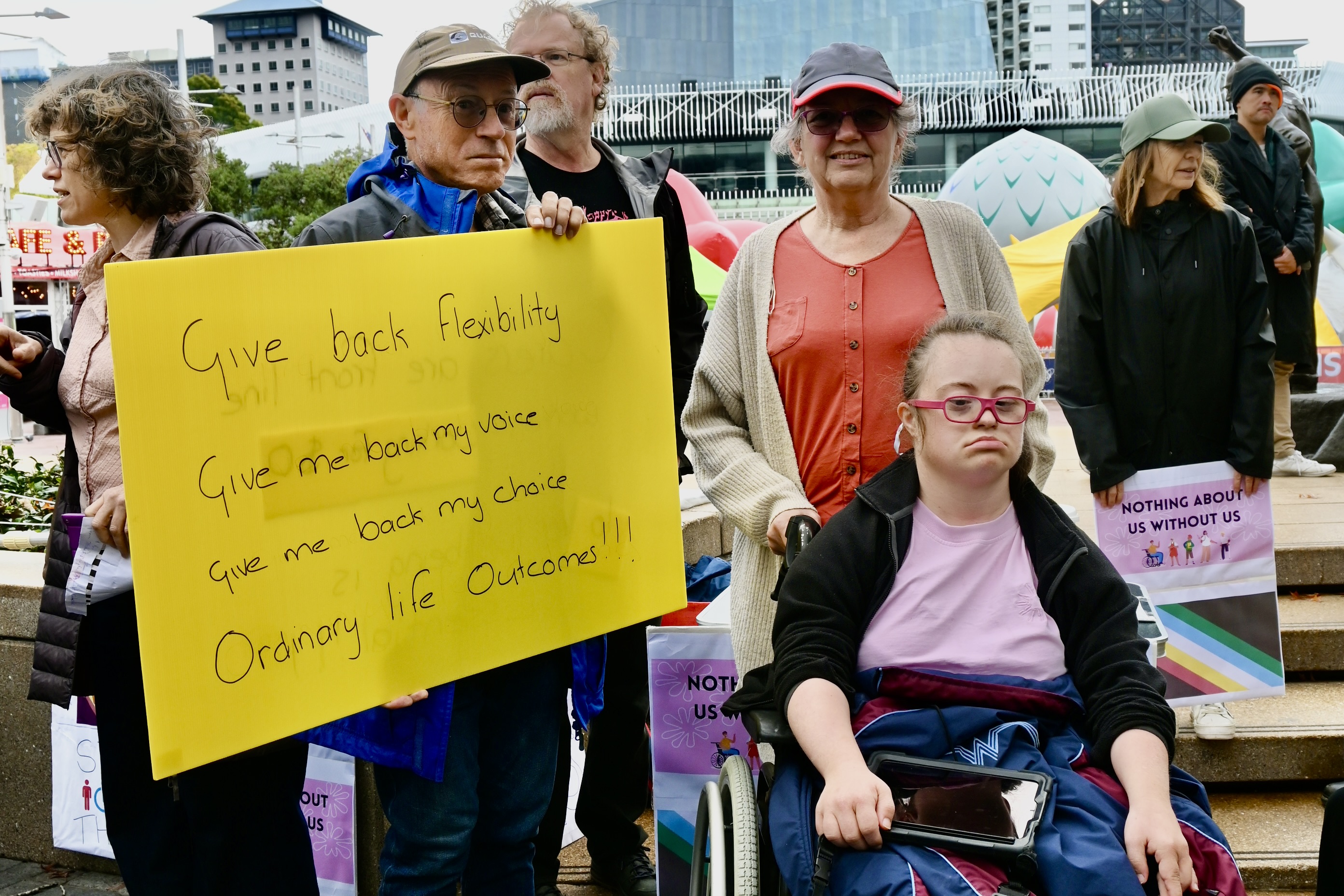 A family with a young woman in a wheelchair hold up a sign that reads: Give back flexibility, give me back my voice, give me back my choice, ordinary life outcomes!!!